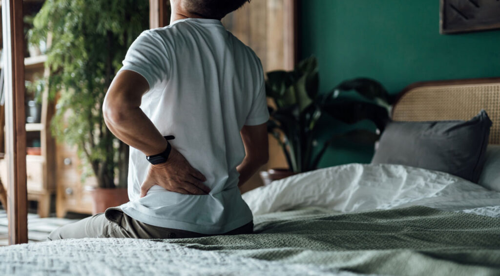 person-sitting-on-the-edge-of-a-bed-holding-their-lower-back person-sitting-on-the-edge-of-a-bed-holding-their-lower-back